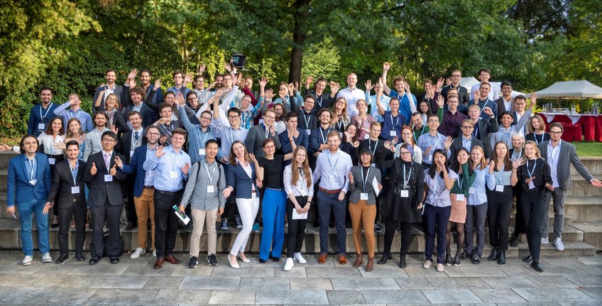 Students of all three Max Planck Schools at Kick-off-symposium of the Max Planck Schools in September 2019. © David Ausserhofer A large group of individuals in professional attire are gathered outdoors, smiling, waving, and posing together on stone steps. Trees and a tent are visible in the background, suggesting a formal gathering or event. © David Ausserhofer