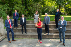 Eine Gruppe von sieben Personen in Business-Kleidung steht auf einer Treppe, umgeben von Bäumen und Grünflächen.