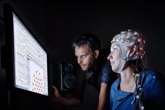 A person is wearing an EEG cap with electrodes while a researcher analyzes data on a screen