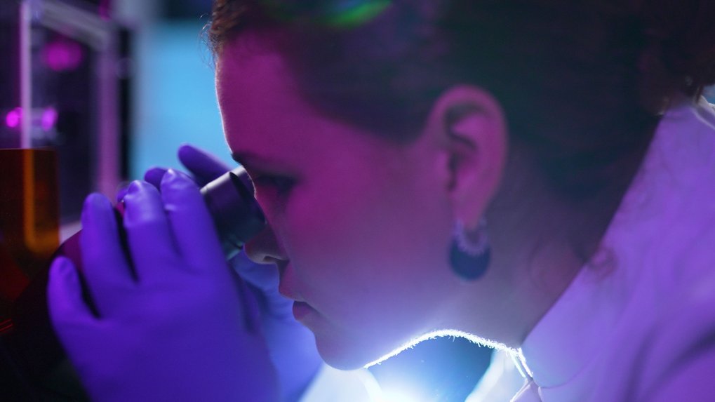 A researcher, wearing gloves and a lab coat, peers into a microscope in a laboratory setting, with purple lighting creating a focused atmosphere.