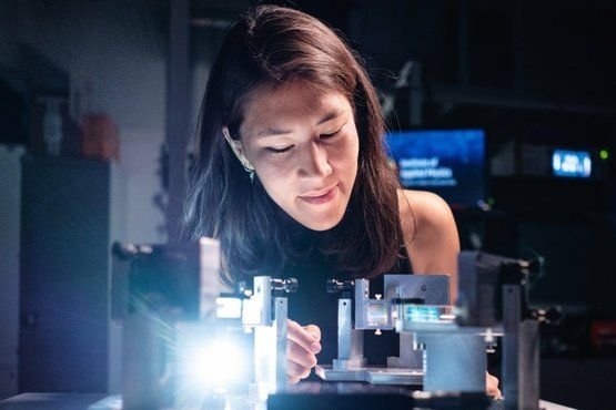 A female scientist is working on a photonics experiment in a lab. She is focused on adjusting small optical components on a table illuminated by a bright light source.
