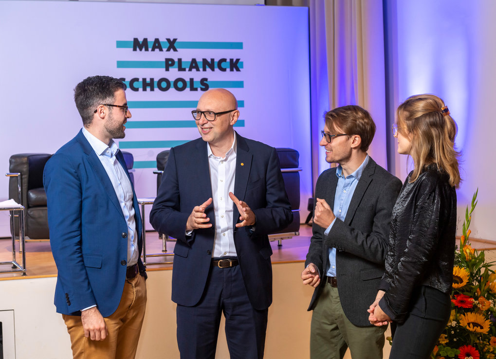 A group of four people, dressed in business attire, engaged in conversation at a Max Planck Schools event, with chairs and floral arrangements in the background.