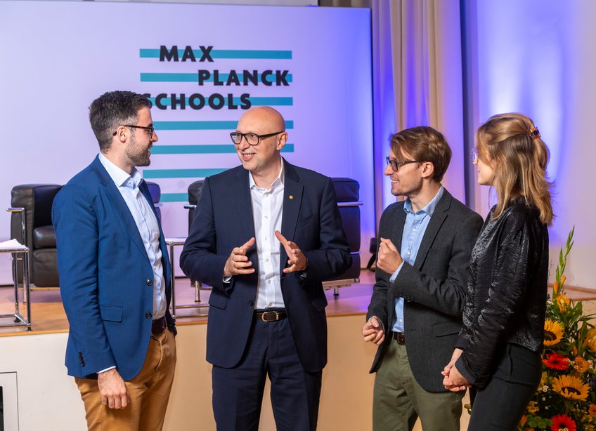 Studierende der Max Planck Schools im Gespräch mit Stefan Hell, Nobelpreisträger und Fellow der Max Planck Schools Matter to Life und Photonics, im Rahmen des Kick-Off-Symposium im September 2019. ©Peter Himsel Vier Personen stehen in einem lebhaften Austausch vor einer Bühne, auf der ein Schild mit der Aufschrift "Max Planck Schools" zu sehen ist. Rechts im Bild befindet sich ein Blumenarrangement.