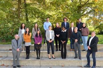 A diverse group of individuals stands on tiered stone steps in a park, featuring lush greenery and autumnal tree foliage in the background.