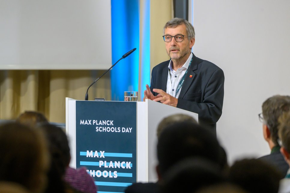 Walter Rosenthal is giving a lecture during Max Planck Schools Day. He is standing at a podium with the "Max Planck Schools" logo. Blurred people can be seen in the background. Rosenthal is wearing a dark suit and glasses and is speaking with gesturing hands.