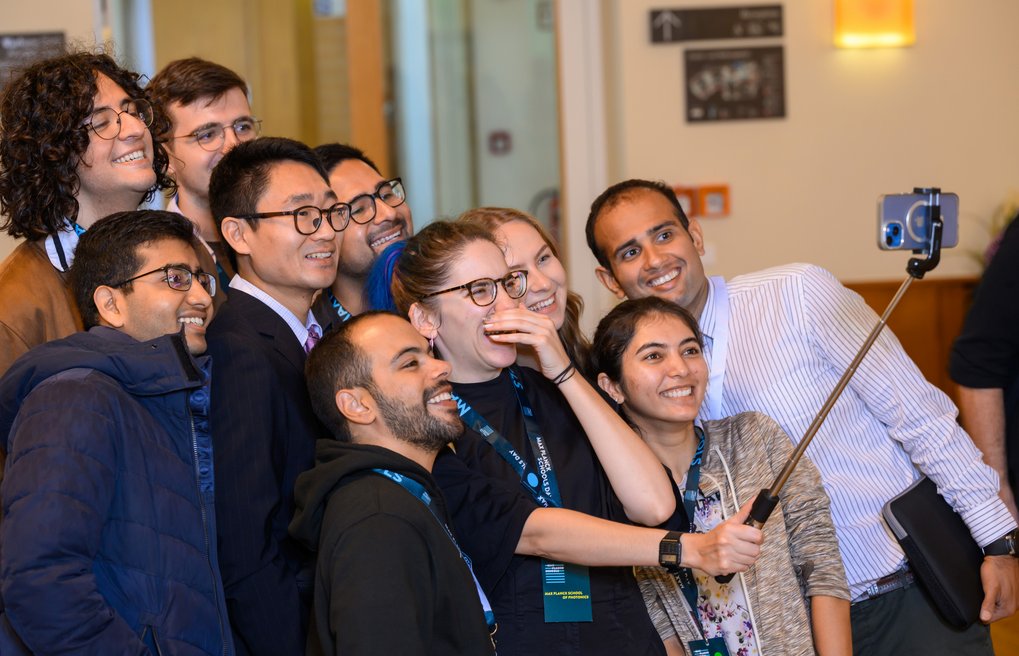 A group of individuals wearing conference lanyards take a selfie together indoors, using a selfie stick to hold the smartphone.