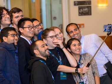 A group of individuals wearing conference lanyards take a selfie together indoors, using a selfie stick to hold the smartphone.