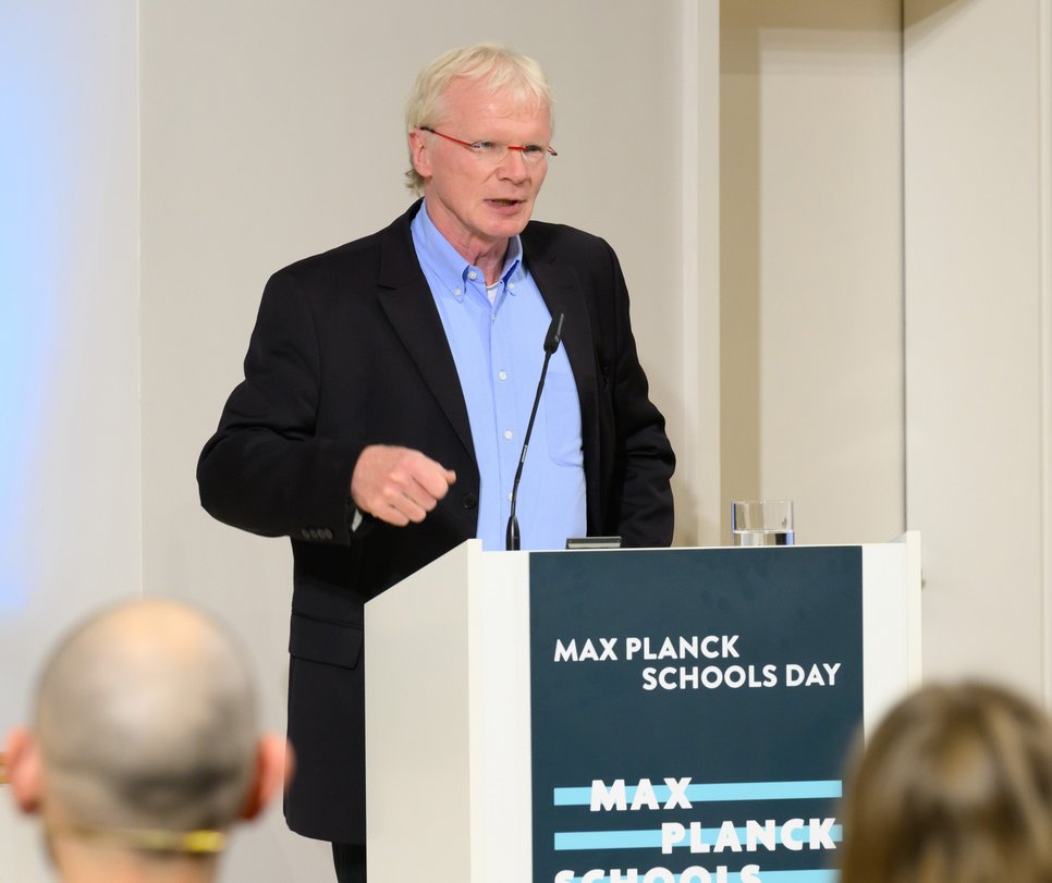 Ferdi Schüth is standing at a lectern and speaking during an event. He is wearing a suit and red glasses. The logo of the Max Planck Schools is on the lectern.