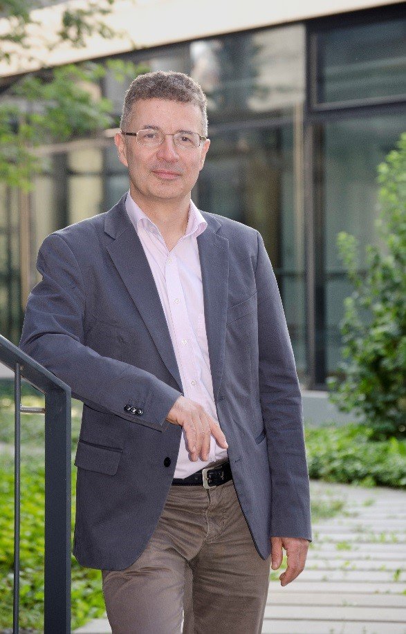 A person dressed in a gray blazer and light pink shirt stands by a railing, in front of a modern glass building with lush greenery.