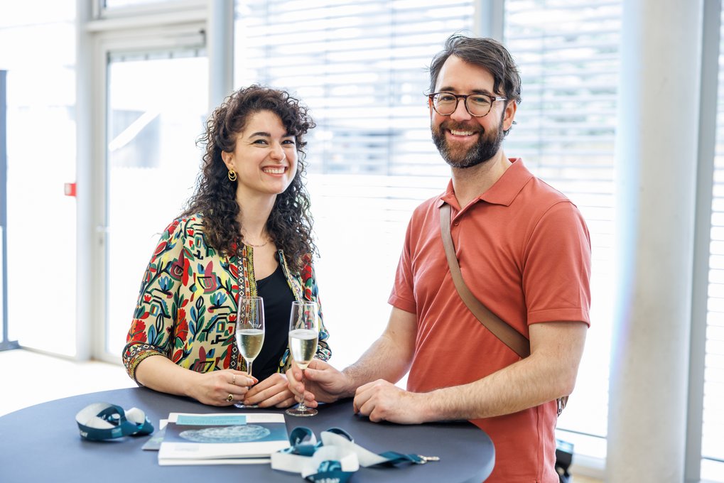 Zwei Personen stehen an einem runden Tisch, halten Sektgläser und sind umgeben von Broschüren und Bändern, die auf dem Tisch liegen.