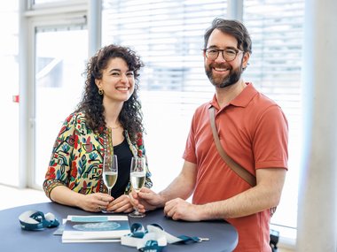 Two individuals engage in conversation at a standing table, each holding a glass of champagne. On the table are lanyards and a book featuring a circular design.
