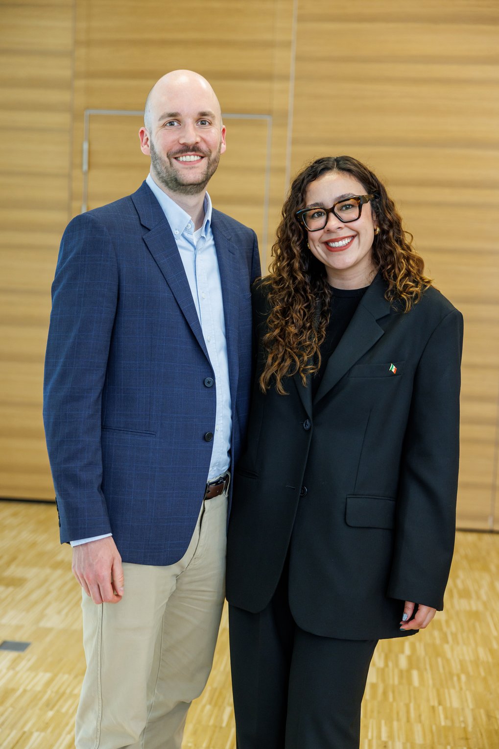 Zwei Personen in formeller Kleidung, eine in einem dunkelblauen Sakko, die andere in einem schwarzen Anzug, stehen nebeneinander auf einem Parkettboden vor einer Holzvertäfelung.