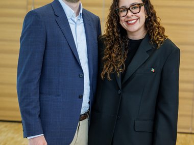 Two individuals in formal attire stand in a room with wooden paneling.