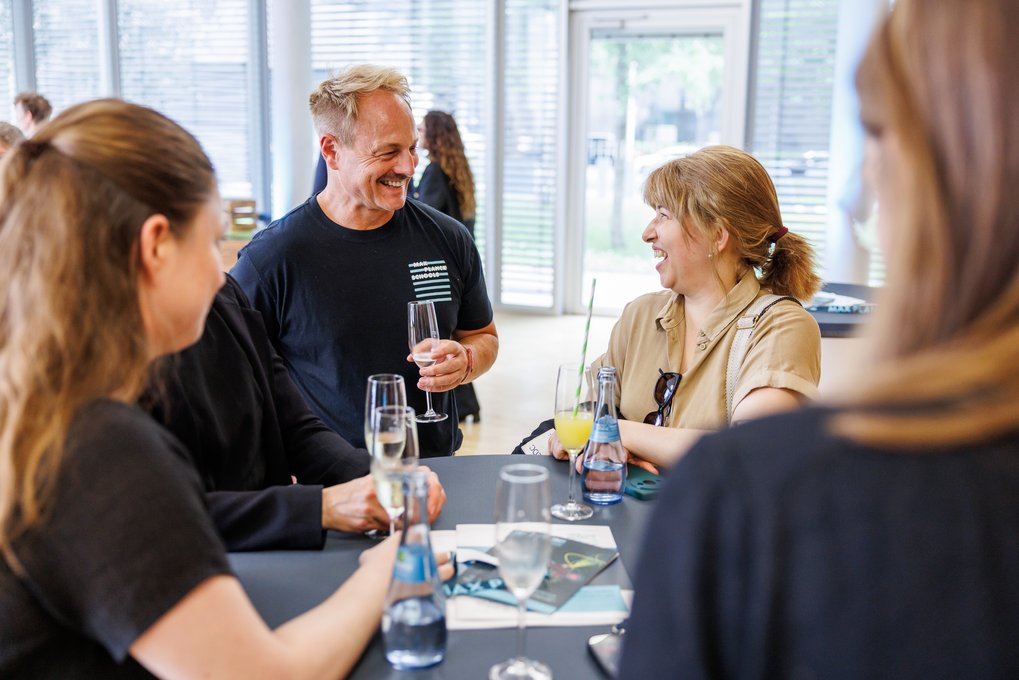 Eine Gruppe von vier Personen steht an einem Tisch bei einem Empfang. Es sind mehrere Getränkeflaschen und Gläser auf dem Tisch zu sehen, sowie Broschüren und ein Handy. Der Raum hat große Fenster im Hintergrund.