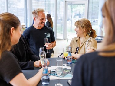Individuals are gathered around a table, holding glasses of drinks and engaging in conversation, in a well-lit, modern space with large windows and visible greenery outside.