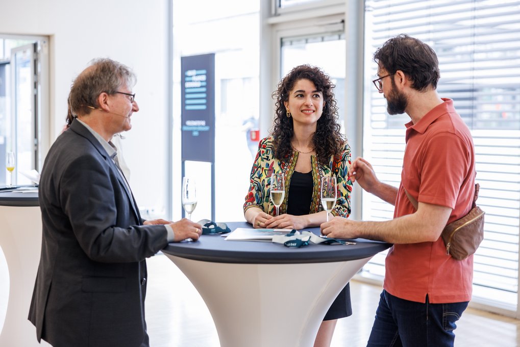 Drei Personen stehen an einem Stehtisch, trinken Sekt und führen ein Gespräch in einem hellen Raum mit großen Fenstern.
