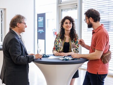 Drei Personen stehen an einem Stehtisch, trinken Sekt und führen ein Gespräch in einem hellen Raum mit großen Fenstern.
