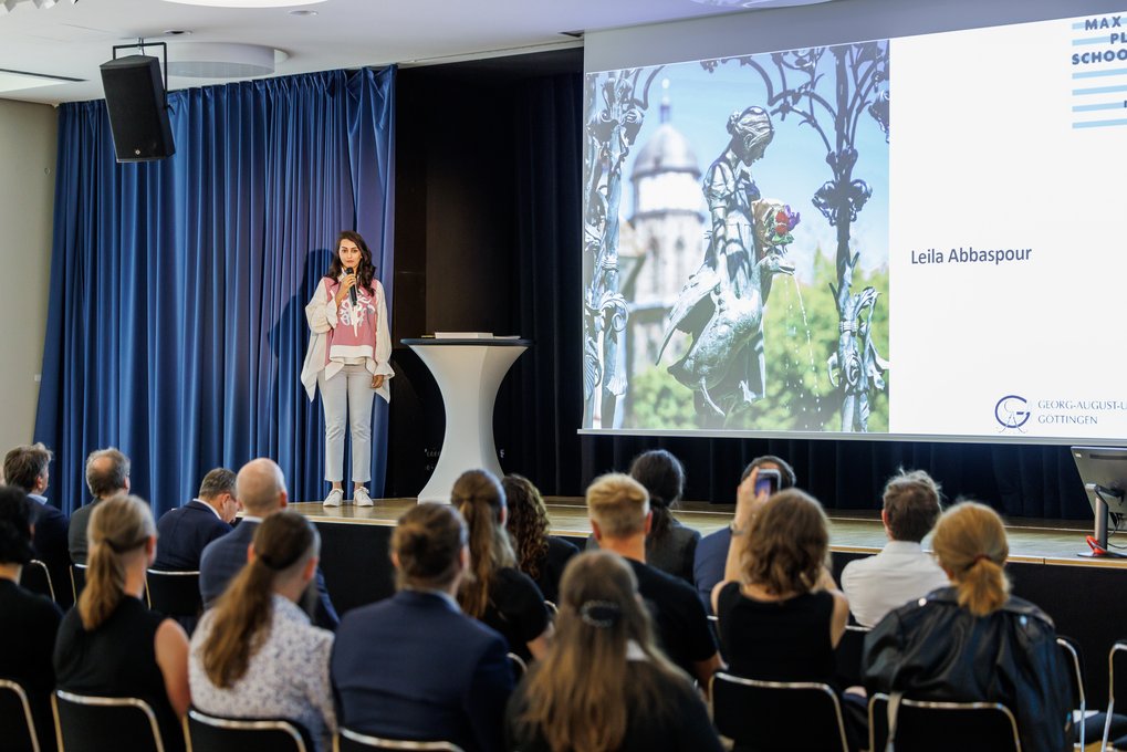 Junge Frau hält Präsentation vor Publikum, steht neben einem Tisch auf einer Bühne, Leinwand zeigt Namen und Bild.