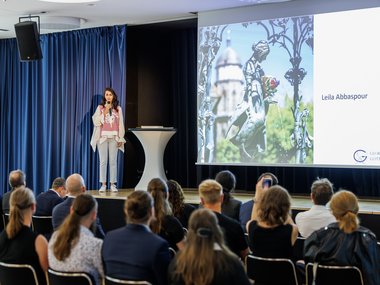Junge Frau hält Präsentation vor Publikum, steht neben einem Tisch auf einer Bühne, Leinwand zeigt Namen und Bild.