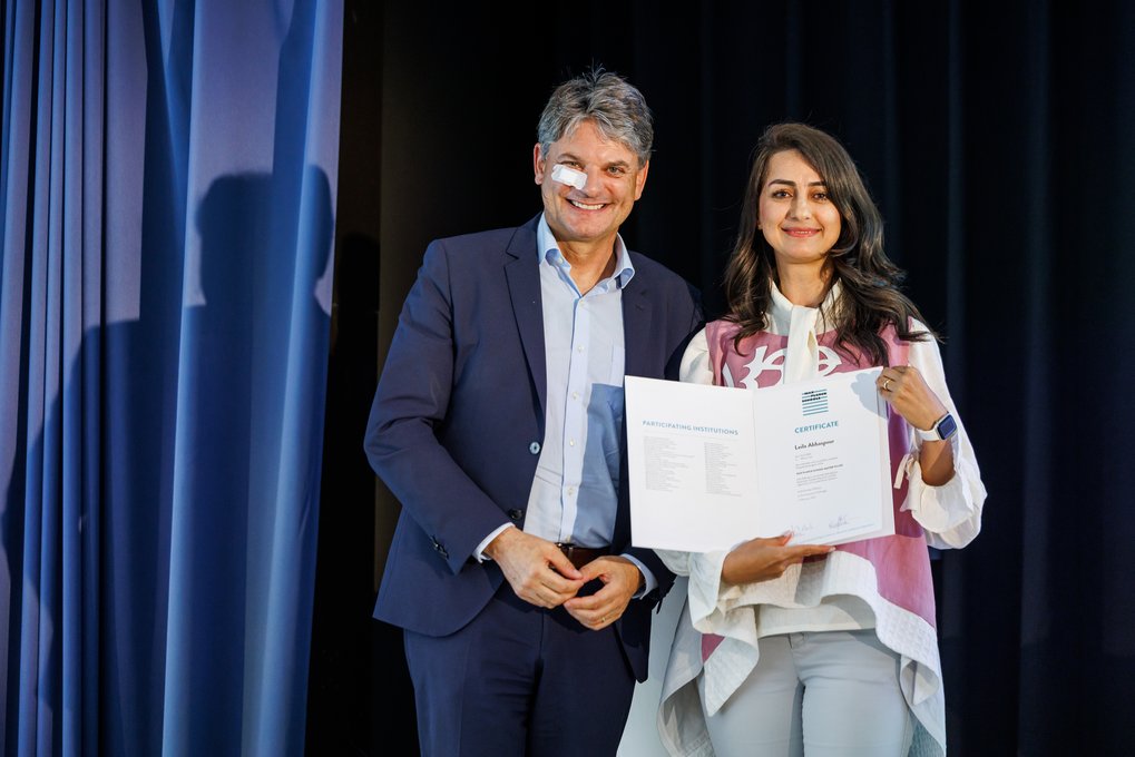 Zwei Personen auf einer Bühne, eine Person hält ein Zertifikat in der Hand.