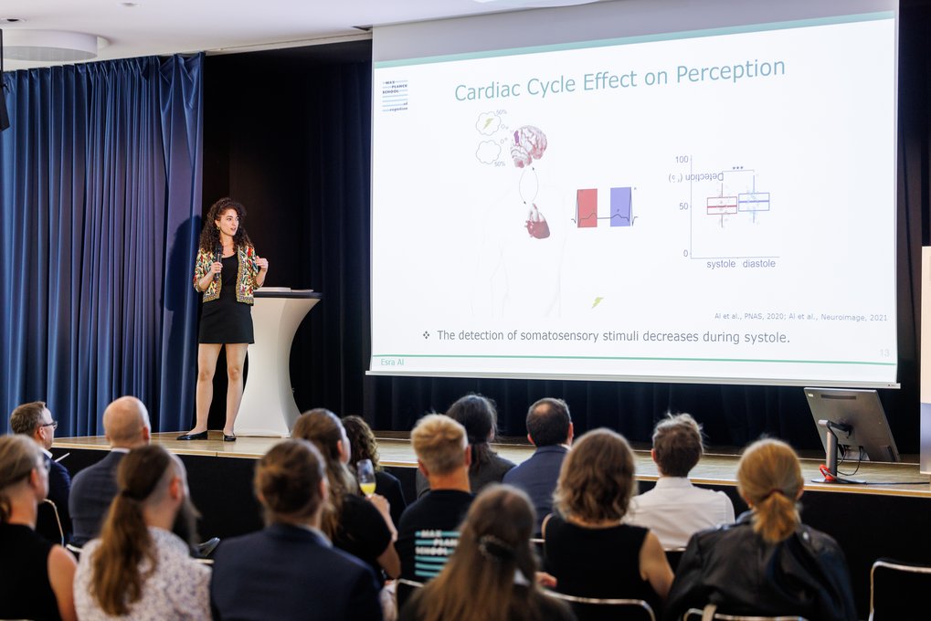 Eine Referentin präsentiert vor Publikum, während eine Folie die Auswirkungen des Herzzyklus auf die Wahrnehmung erklärt und Diagramme zeigt. Zuschauer verfolgen aufmerksam die Präsentation.