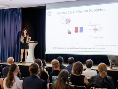A speaker presents to an audience while a slide explains the effects of the cardiac cycle on perception and shows diagrams. Audience members follow the presentation attentively.