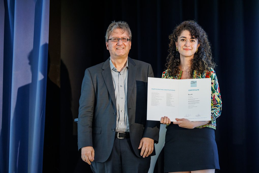 Zwei Personen stehen auf einer Bühne; eine trägt ein buntes Oberteil und hält ein Zertifikat mit englischem Text.