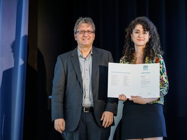 Two people are standing on a stage; one is wearing a colorful top and holding a certificate with English text.