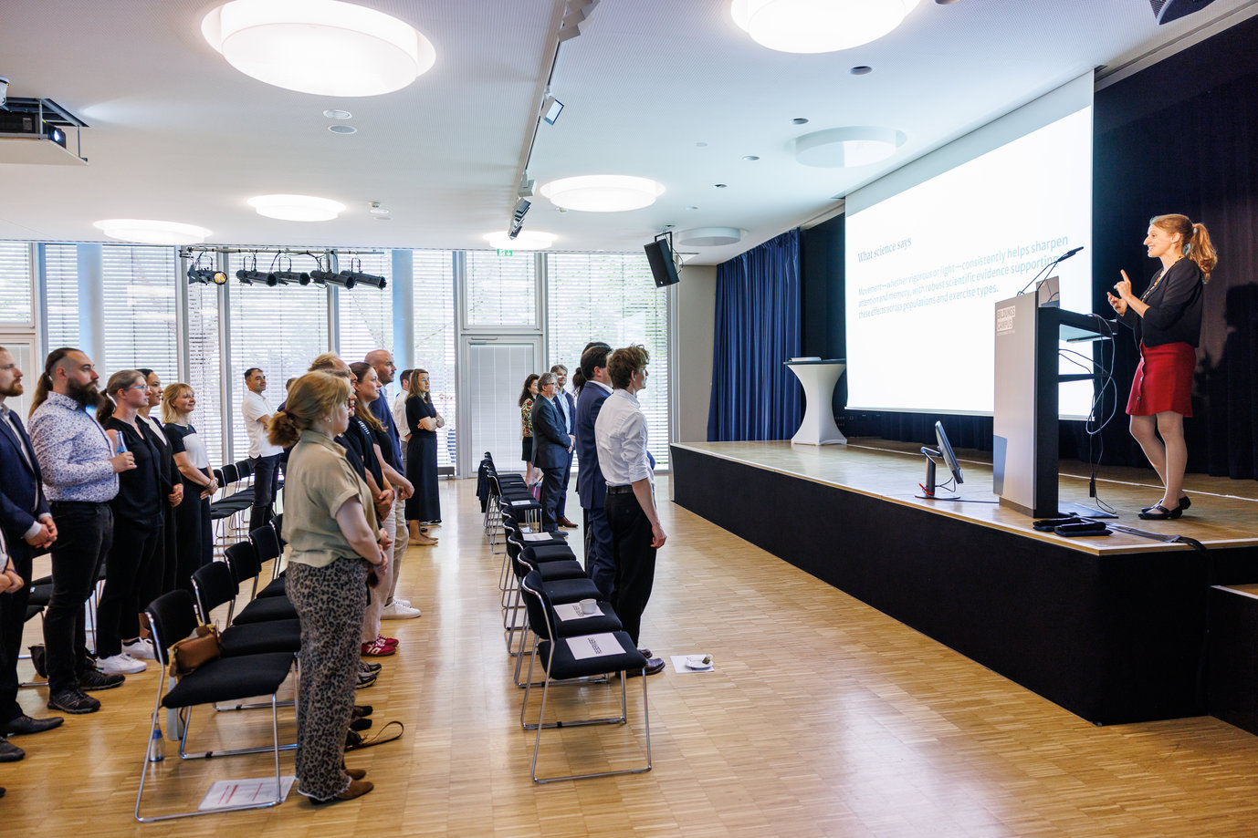 Attendees in a modern conference room stand in rows, watching a presentation on a large screen, with natural light filtering through large windows.