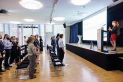 Attendees in a modern conference room stand in rows, watching a presentation on a large screen, with natural light filtering through large windows.