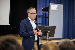 A speaker in a blue blazer and white shirt holds a microphone, addressing an audience at an educational event, with a lectern and screen in the background.