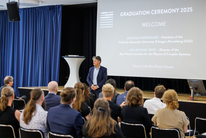 Prof. Joachim Hornegger, ebenso Dean der Max Planck Schools und Präsident der FAU Erlangen-Nürnberg, richtete sehr persönliche Worte an die Alumni vor Ort Graduierungszeremonie 2025 mit Publikum und Redner vor Projektorbildschirm.