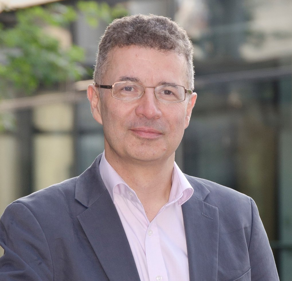 Professional middle-aged man with short curly hair and glasses, dressed in a suit jacket and light pink shirt, posing outdoors in front of a building.