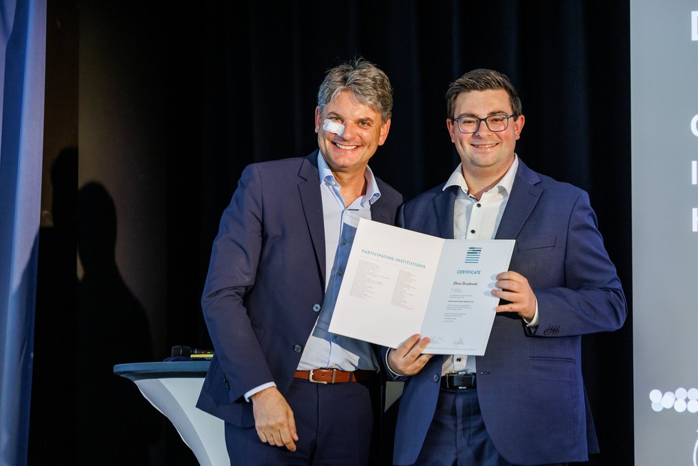 Zwei Männer in dunklen Anzügen stehen nebeneinander und präsentieren ein geöffnetes Zertifikat auf einer Bühne vor einem dunklen Hintergrund.