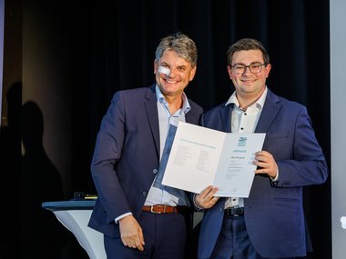 Two men in dark suits stand side by side and present an open certificate on a stage in front of a dark background.