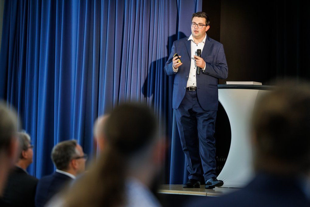 Ein Mann in einem blauen Anzug steht auf einer Bühne mit einem Mikrofon und einem Präsentationsgerät.