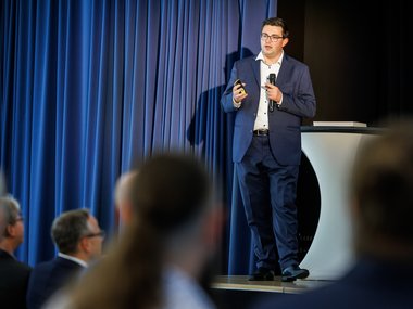 Ein Mann in einem blauen Anzug steht auf einer Bühne mit einem Mikrofon und einem Präsentationsgerät.