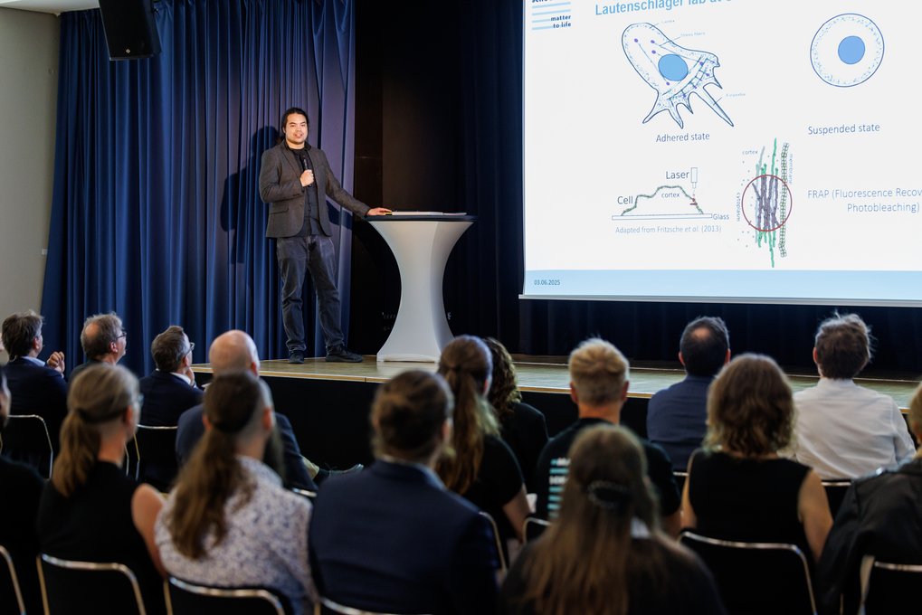 Wissenschaftlicher Vortrag über Zellzustände, Diagramme auf großer Leinwand, Publikum hört interessiert zu. Bühne mit blauem Vorhang.
