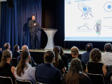 Scientific lecture on cell states, diagrams on a large screen, audience listening intently. Stage with blue curtain.