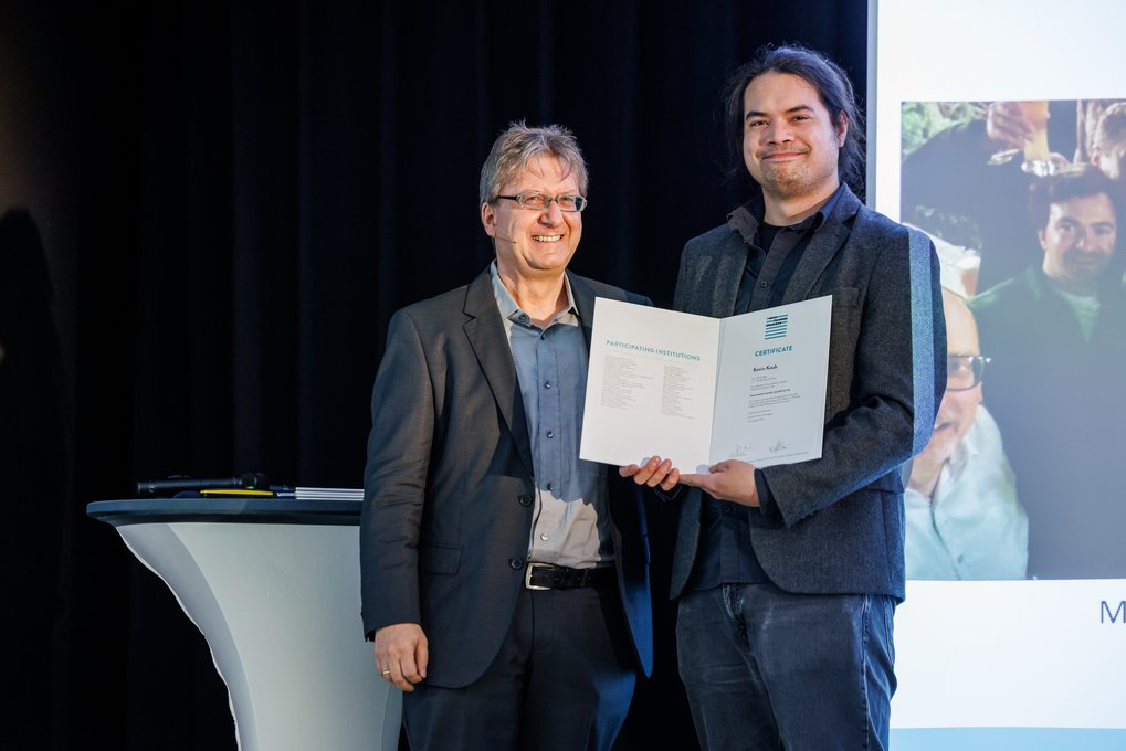 Zwei Personen in Anzügen halten ein Zertifikat auf einer Bühne. Neben ihnen steht ein Tisch mit Stiften. Im Hintergrund ist ein Bildschirm sichtbar.