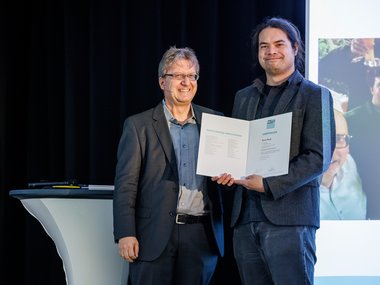 Two people in suits are holding a certificate on a stage. Next to them is a table with pens. A screen is visible in the background.