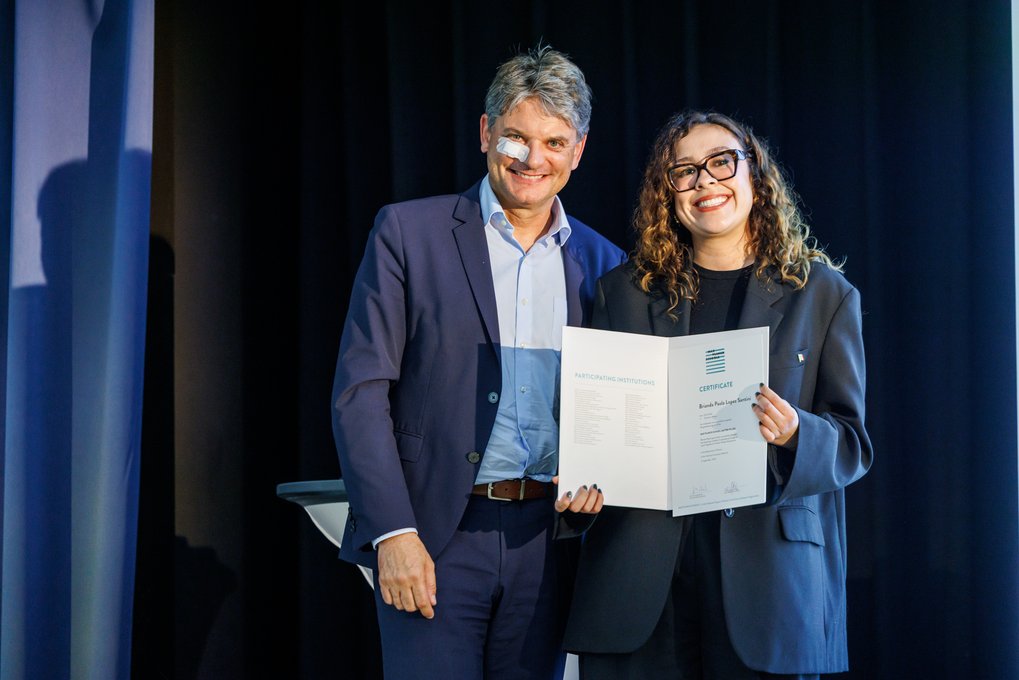 Zwei Personen in formeller Kleidung präsentieren ein Zertifikat vor einem dunklen Hintergrund, möglicherweise bei einer Preisverleihung oder einem offiziellen Ereignis.