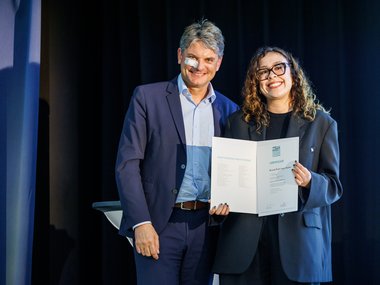 Two people in formal attire present a certificate in front of a dark background, possibly at an award ceremony or official event.