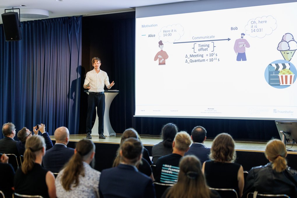 Vortragender präsentiert technische Inhalte zu Zeitversatz in Kommunikation, Publikum sitzt in einem Konferenzraum. Diagramm zeigt Figuren Alice und Bob mit Kommunikatonszeit.