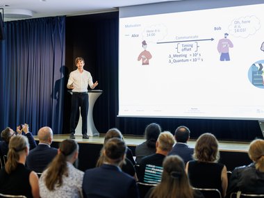 Speaker presents technical content on time lag in communication, audience sits in a conference room. Diagram shows characters Alice and Bob with communication time.
