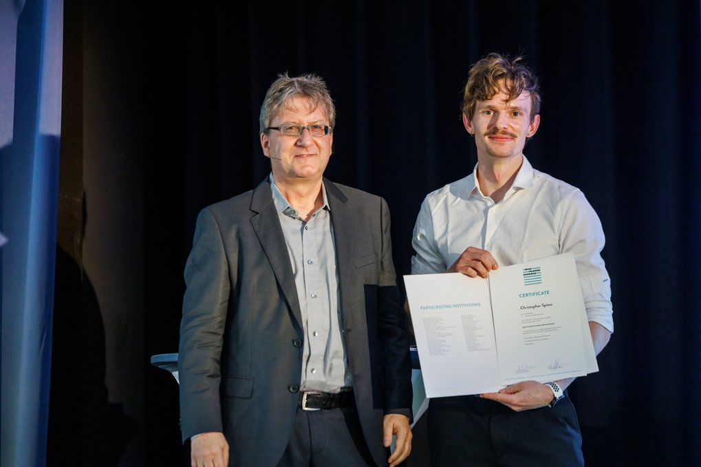 Zwei Männer stehen auf einer Bühne. Der Mann rechts präsentiert ein Zertifikat mit Text und Logos darauf. Sie tragen formelle Kleidung.