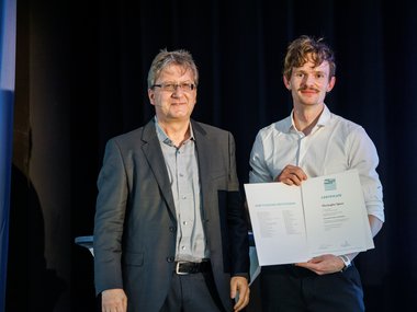 Two men are standing on a stage. The man on the right is presenting a certificate with text and logos on it. They are wearing formal attire.