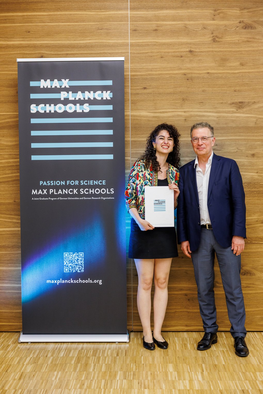 Zwei Personen posieren vor einem Banner der Max-Planck-Schools, eine hält ein Zertifikat. Das Banner zeigt den Slogan "Passion for Science" und enthält eine Webadresse.