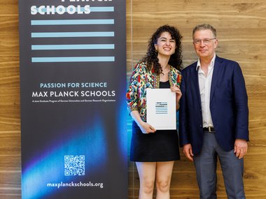 Zwei Personen posieren vor einem Banner der Max-Planck-Schools, eine hält ein Zertifikat. Das Banner zeigt den Slogan "Passion for Science" und enthält eine Webadresse.