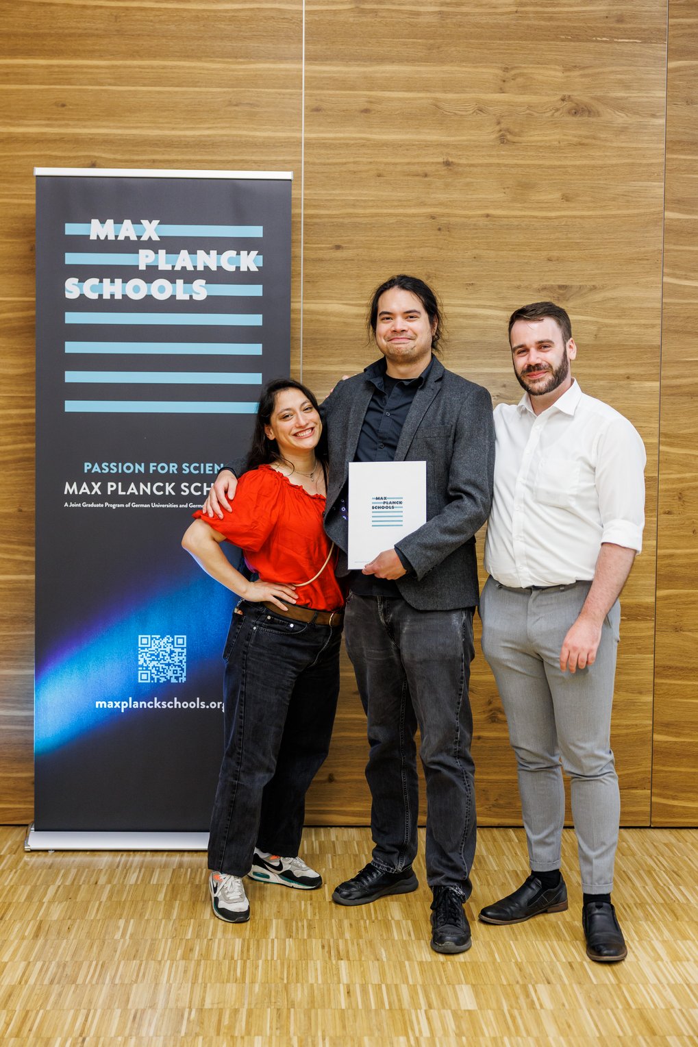 Drei Personen stehen vor einem Max Planck Schools Banner.